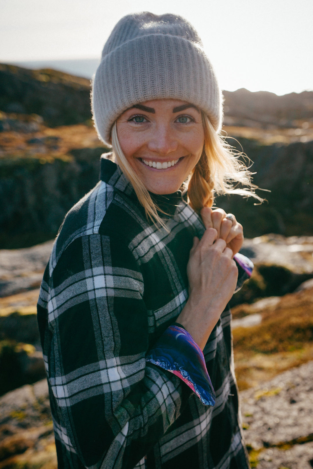 Smiling woman wears Pladra’s Women’s Northwood Sherpa Insulated Flannel Jacket in RP Onyx Black among sunlit rocks.