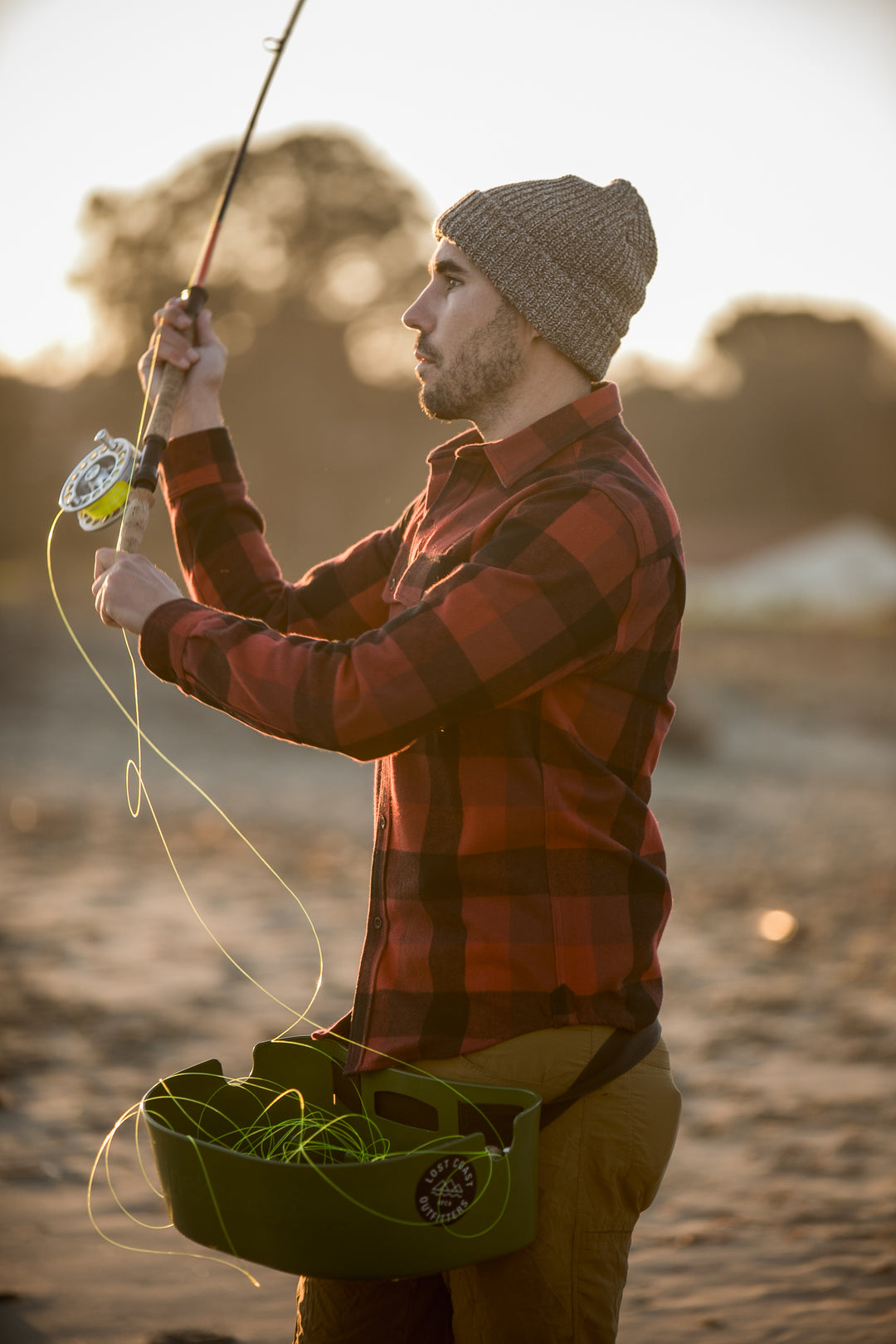 A man in a plaid shirt and knit hat fly fishing on a sandy riverbank at sunset.