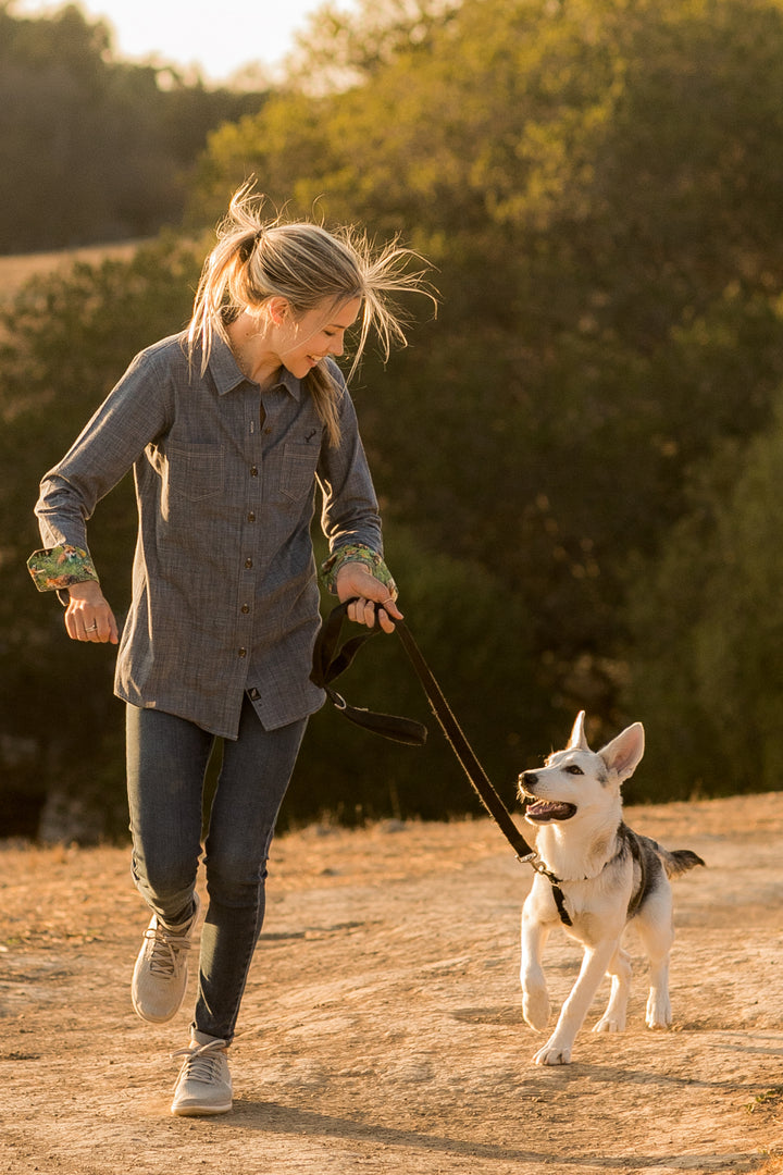 A woman with blonde hair jogging on a dirt path at sunset, smiling at her dog on a leash. She wears the Women&