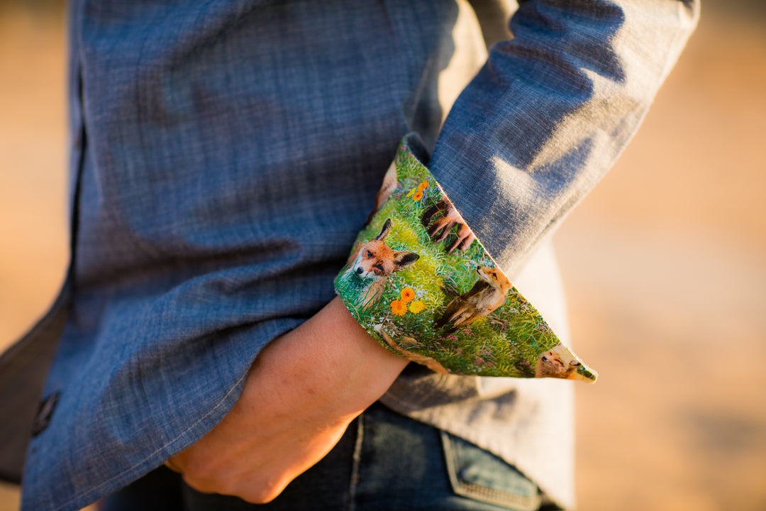 A person wears a gray jacket with cuffs turned up to show a fox and green forest print lining. Underneath, they have the Women&