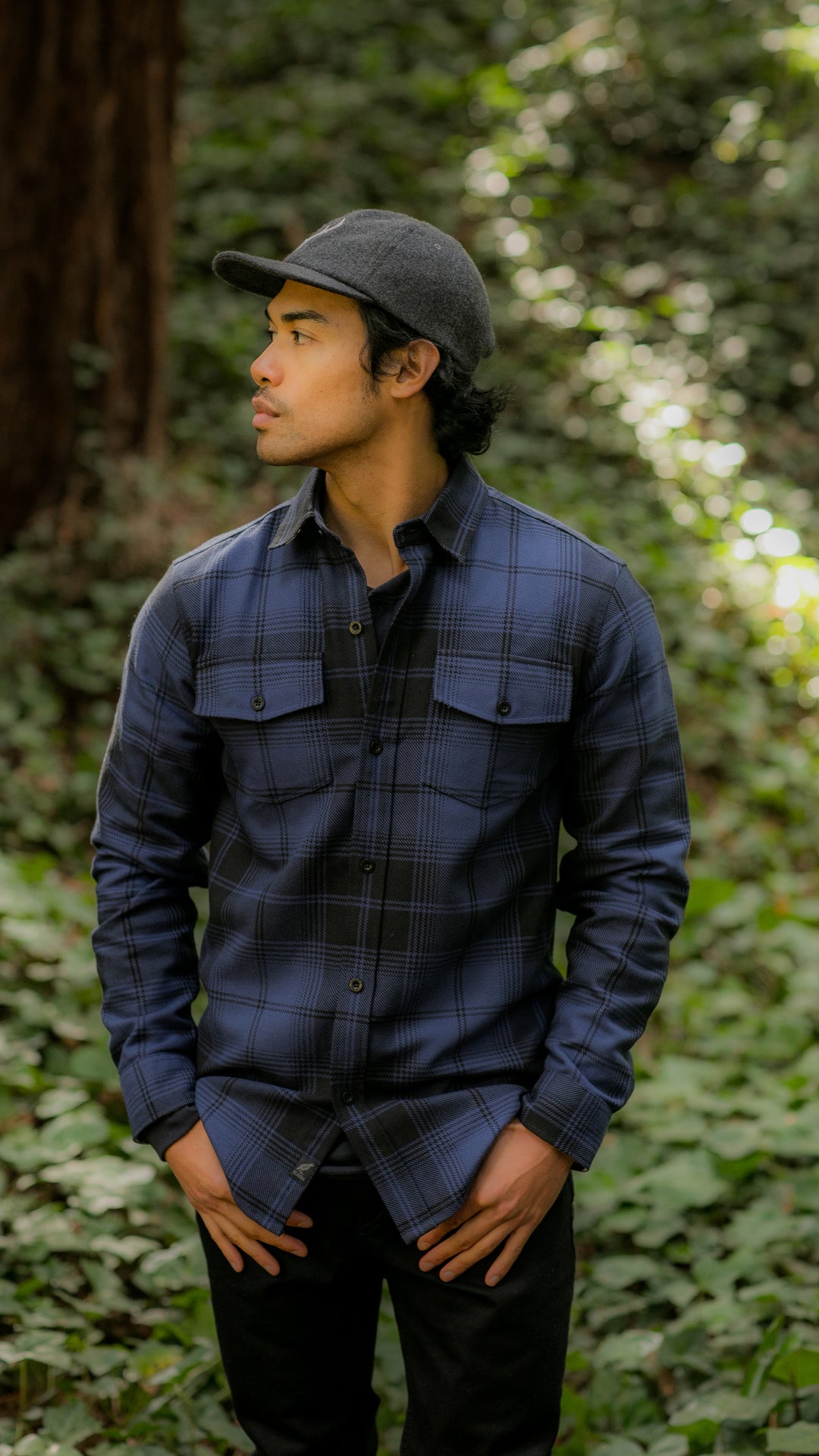 Man in a blue plaid shirt and gray cap stands in a forested area, looking to the side with hands in pockets.