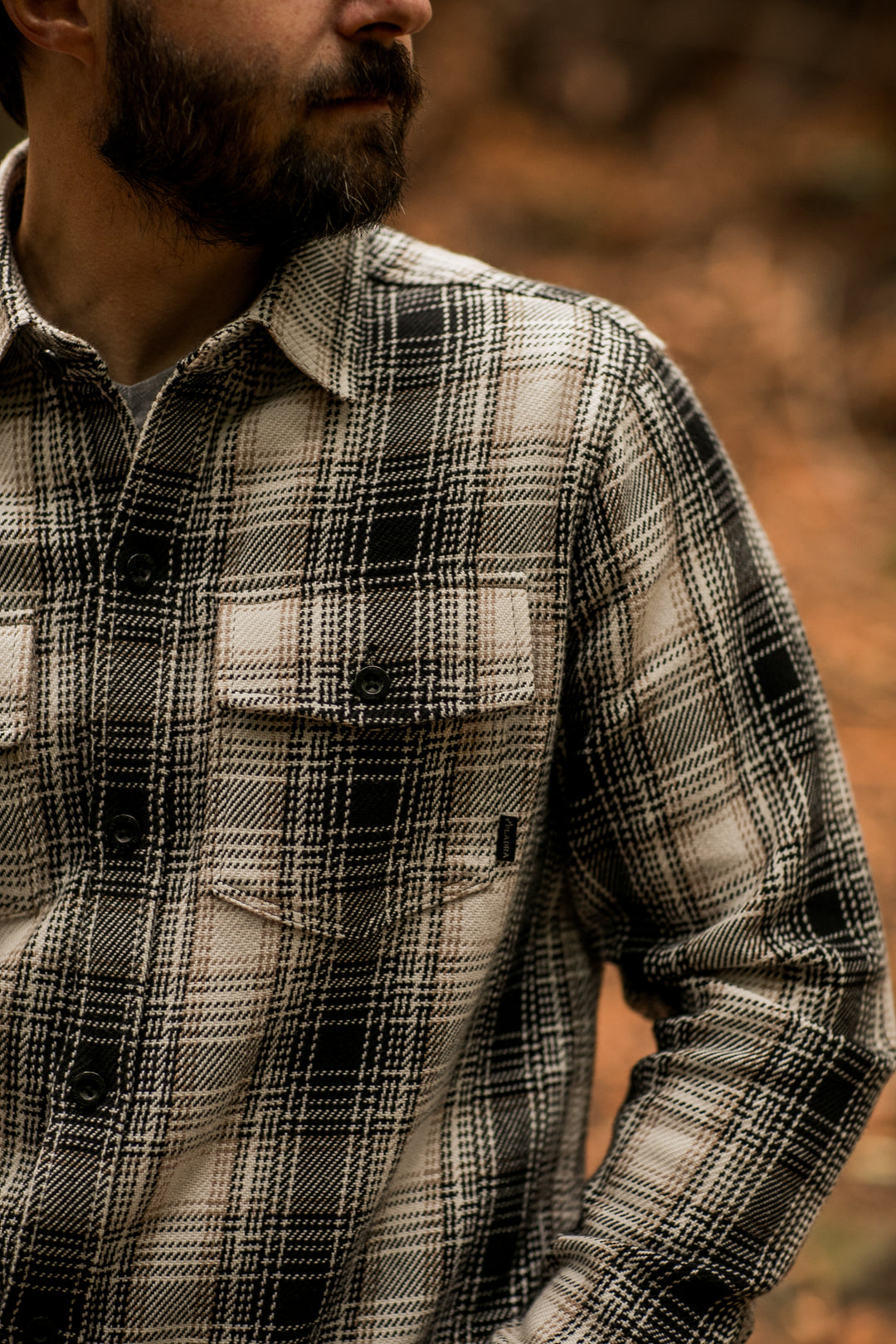 A man with a beard wears a black and white plaid shirt, standing outdoors with a blurred brown background.