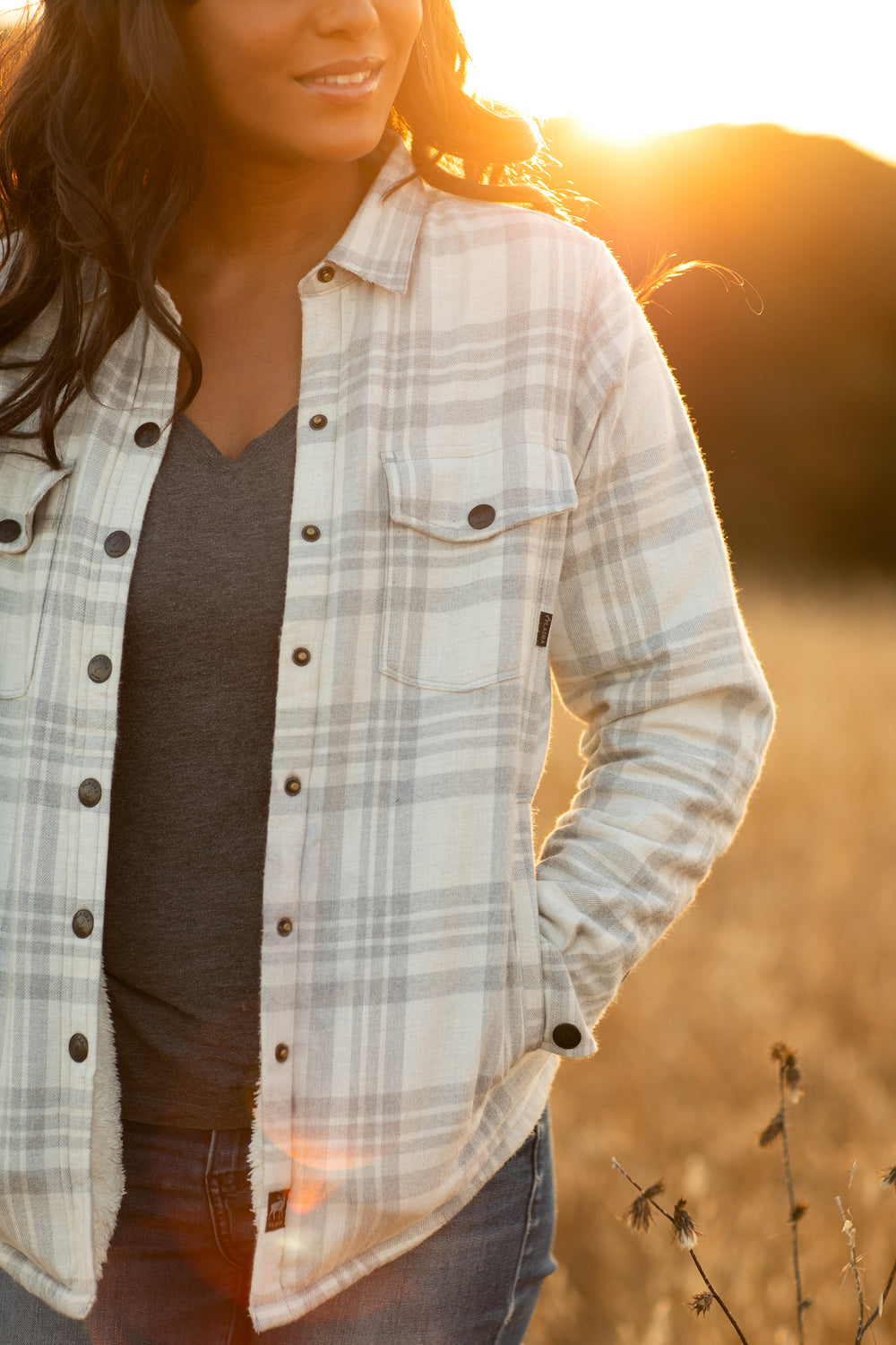 A woman in the Pladra Women's Northwood Sherpa Insulated Flannel Jacket - Cloud Grey stands in a sunlit field, hands in pockets.