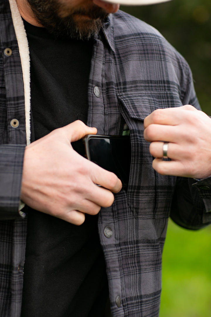 A person slips a black smartphone into the chest pocket of a Pladra Northwood Sherpa Insulated Flannel Jacket outdoors.