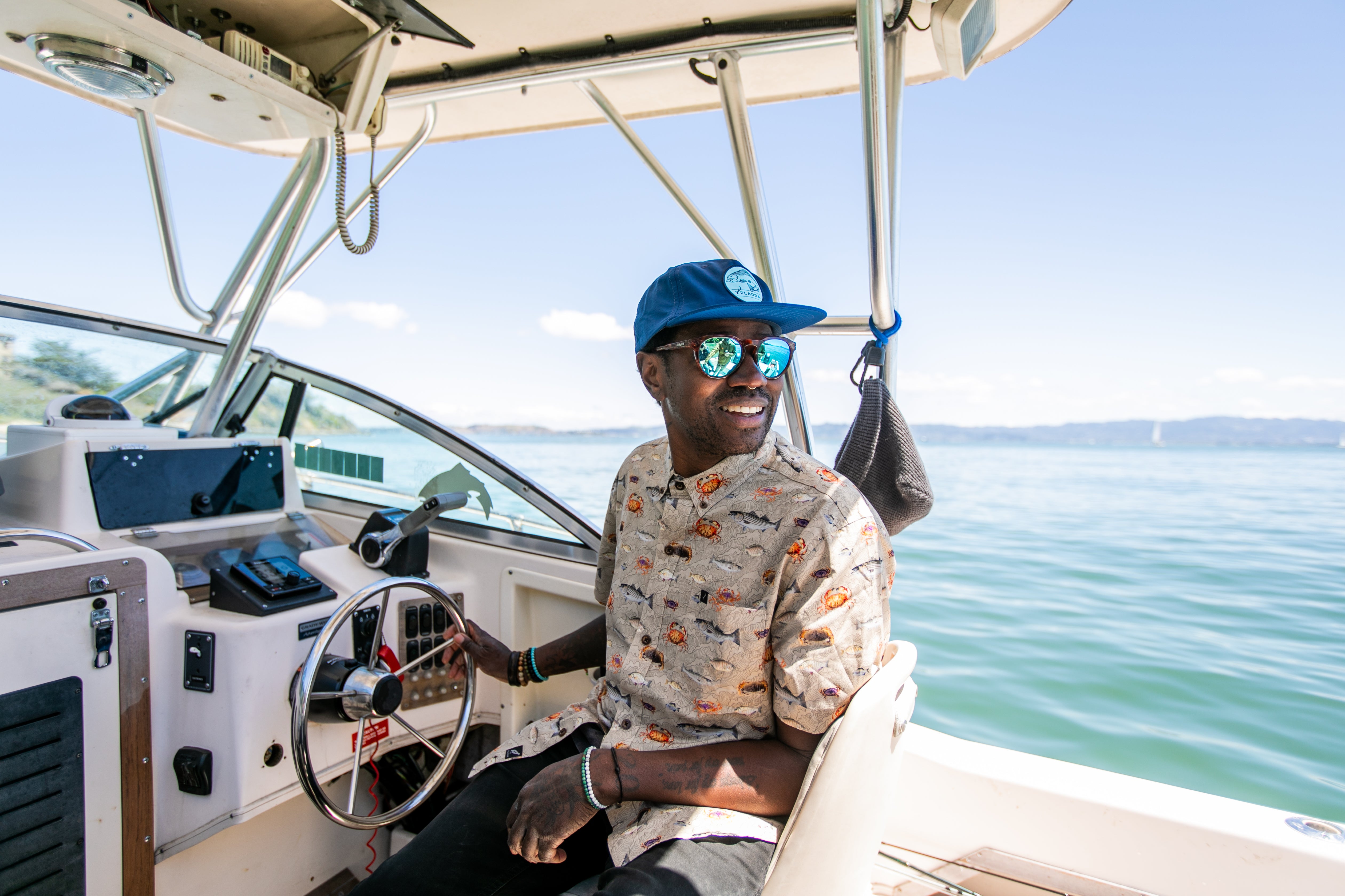 Man in sunglasses and a blue cap steering a boat on a sunny day, smiling, with water and sky in the background.