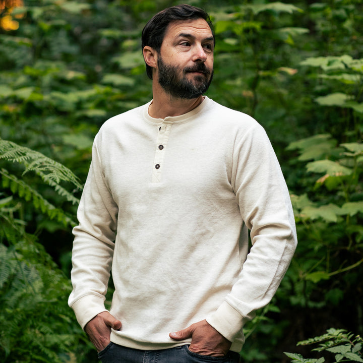 A bearded man stands outdoors before green foliage, hands in pockets, wearing jeans and the Men&