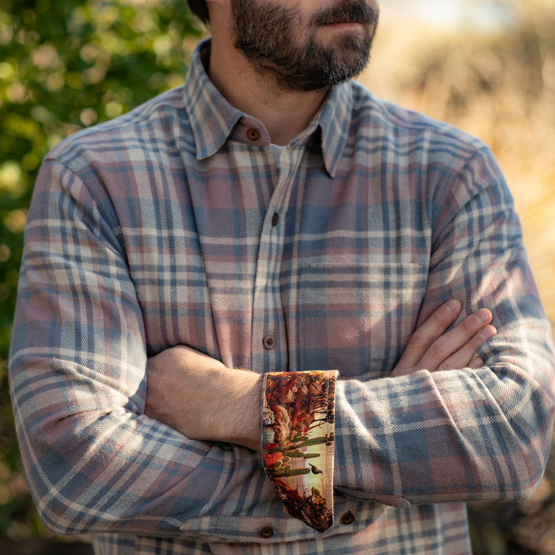 A man in a plaid shirt stands outdoors with arms crossed, showing a colorful patterned shirt cuff.
