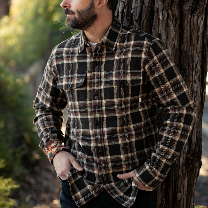 A bearded man stands outdoors by a tree, wearing the Men&