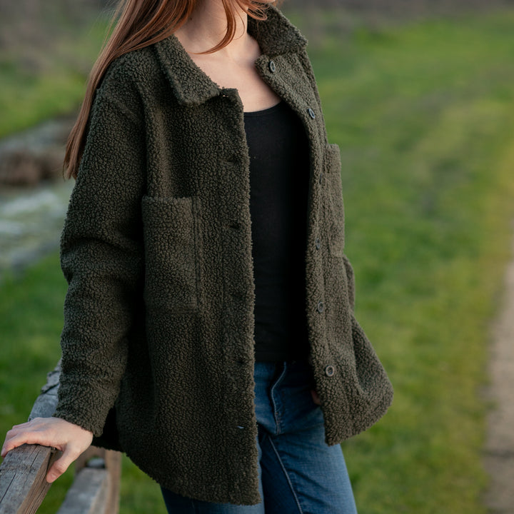 A woman in a Pladra Women's Headlands Sherpa Shacket—Forest Green—stands outdoors, hand on a wooden railing.