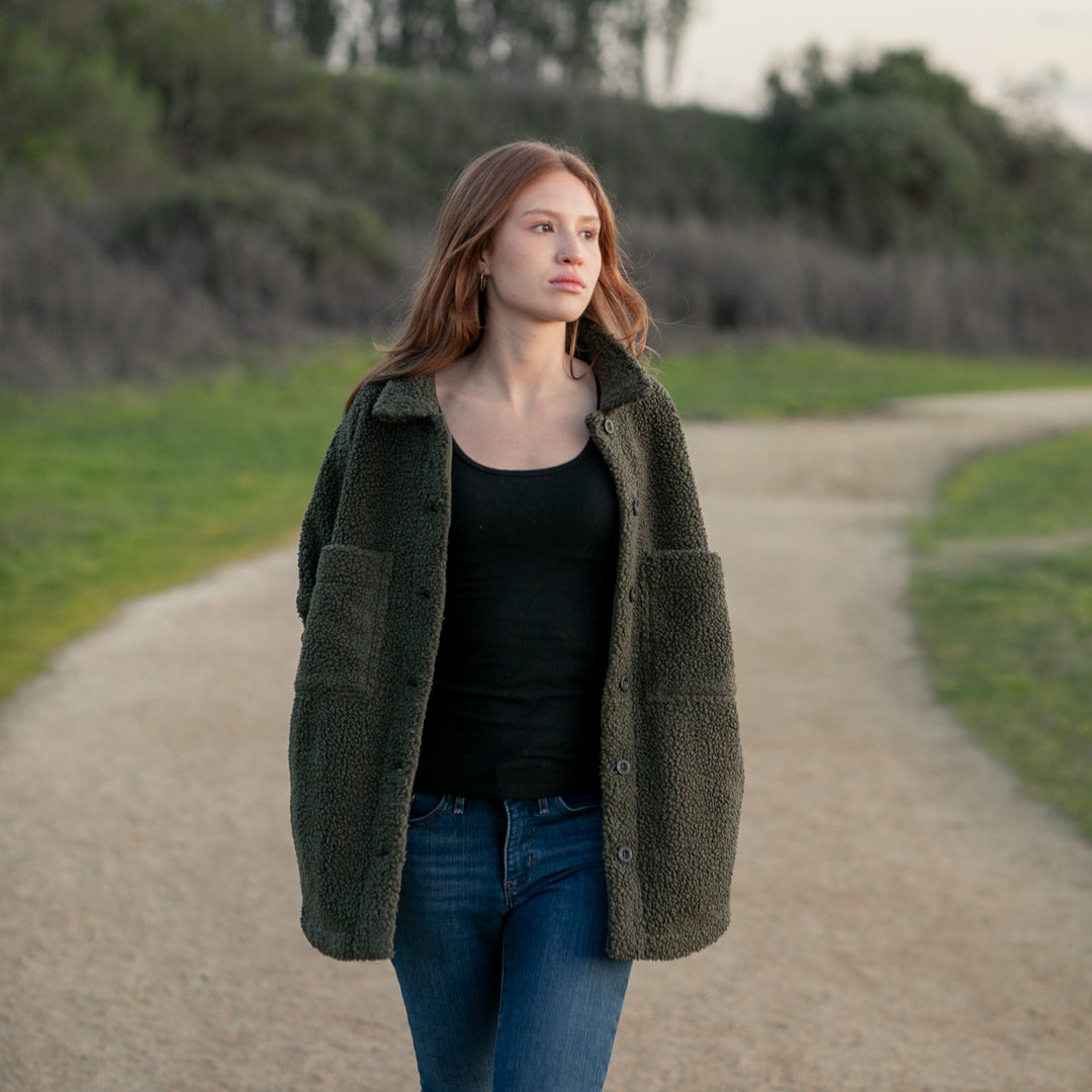 A woman with long hair walks outdoors in a Pladra Women's Headlands Sherpa Shacket- Forest Green, black top, and jeans.