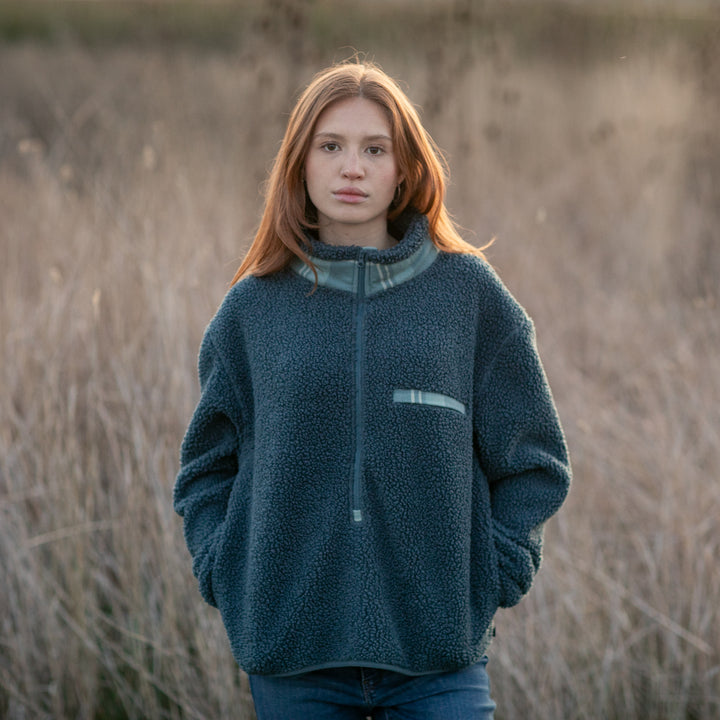 A young woman with long red hair stands in a field of tall grass, wearing the Women&