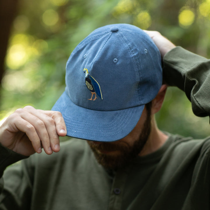 A man in a green shirt adjusts his Pladra Quail Corduroy Hat- Blue with an embroidered bird, outdoors among greenery.