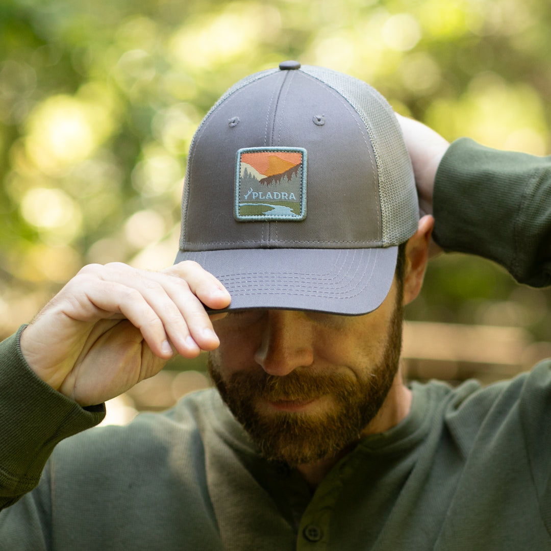 A bearded man adjusts a Mountain Trucker Hat- Charcoal Grey featuring a colorful mountain patch labeled PLADRA. He wears a green long-sleeve shirt outdoors against a leafy, blurred background.
