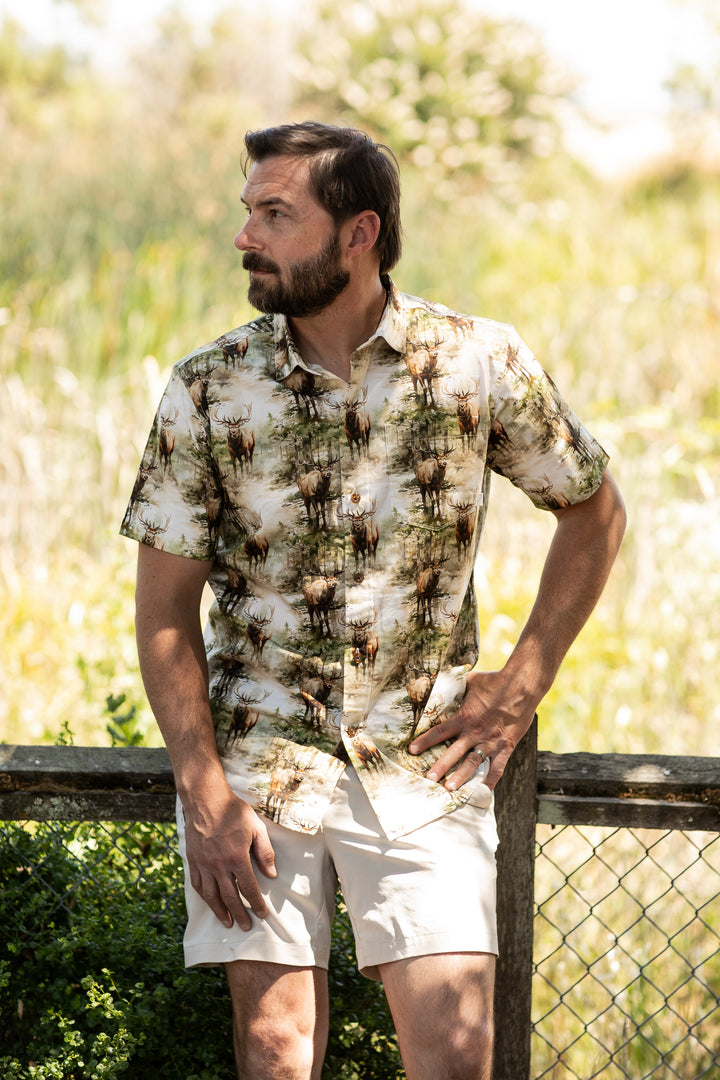 A man with short dark hair and a beard stands by a wooden fence outdoors, wearing the Men&