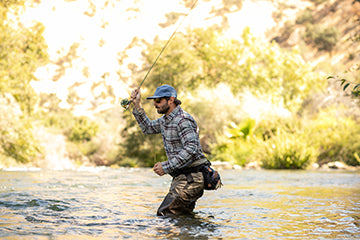 Man in plaid shirt and hat fly-fishing in a shallow river, surrounded by trees and sunlight.