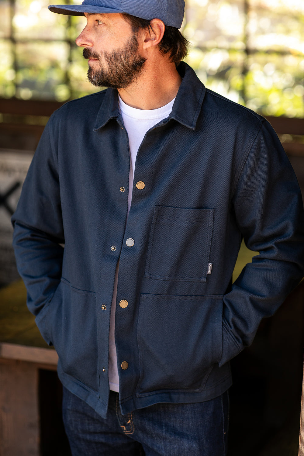 A bearded man in a blue cap, wearing the Men's Sequoia Chore Coat in Midnight Blue with large front pockets and blue jeans, stands with his hands in his pockets, looking to the side.