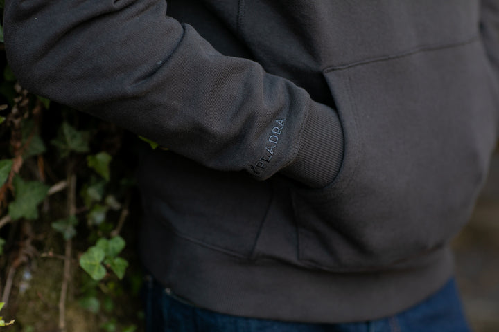 A man in the Pladra Men's Sierra Hoodie - Charcoal Black stands by ivy with a hand in his pocket.