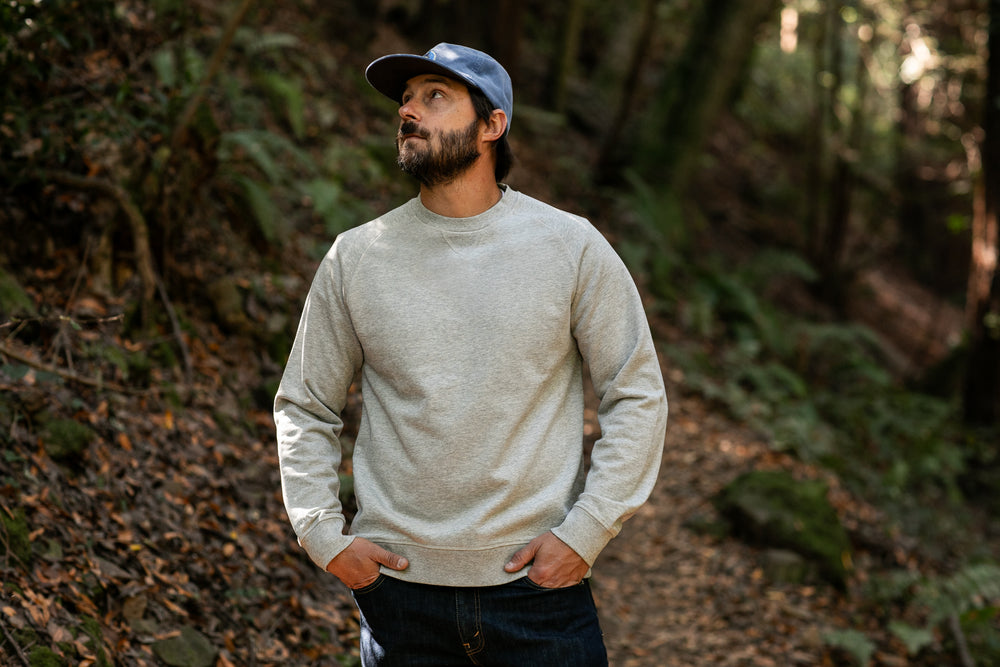 A man wearing the Men's Klamath Crew in Fog Grey and blue jeans stands on a wooded trail with his hands in his pockets, gazing up at the trees as sunlight filters through the forest.