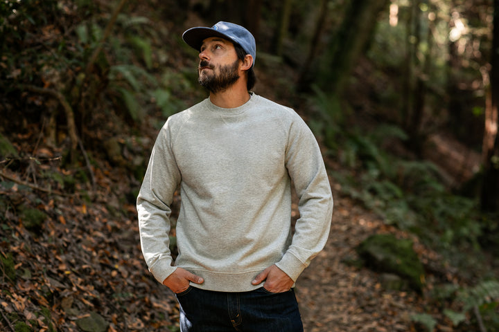 A man wearing the Men's Klamath Crew in Fog Grey and blue jeans stands on a wooded trail with his hands in his pockets, gazing up at the trees as sunlight filters through the forest.