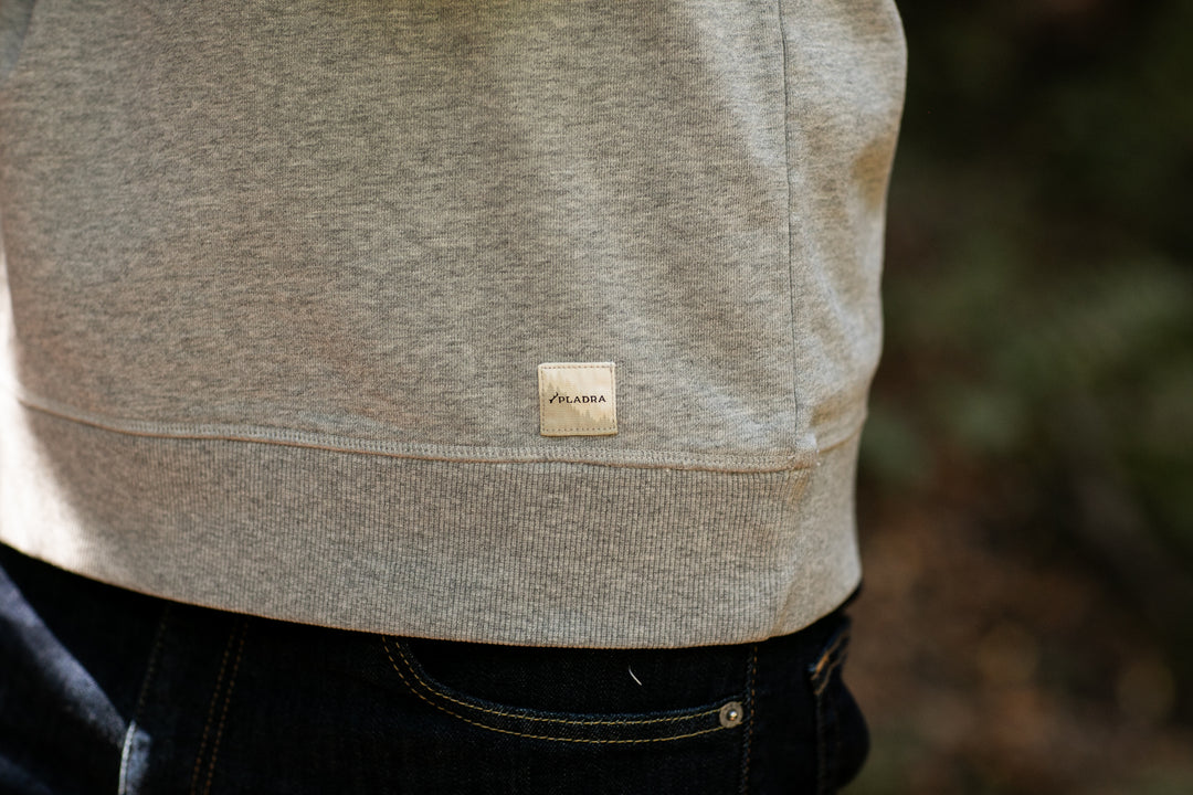 Close-up of the Men's Klamath Crew in Fog Grey, a premium cotton crewneck with a small rectangular VLADA label near the hem. The classic fit is styled with dark pants, set outdoors against blurred greenery.