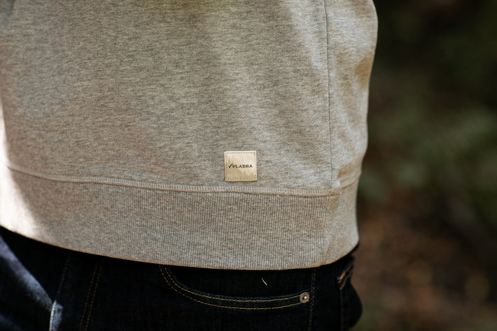 Close-up of the Men's Klamath Crew in Fog Grey, a premium cotton crewneck with a small rectangular VLADA label near the hem. The classic fit is styled with dark pants, set outdoors against blurred greenery.