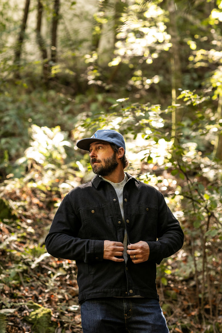A man in jeans and a blue cap stands in a sunlit forest, buttoning his Men&