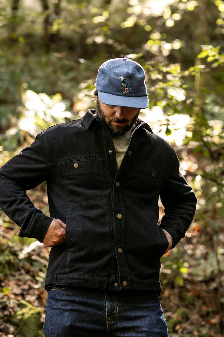 A man in jeans and a blue baseball cap stands outdoors in a sunlit forest, looking down with his hands in the pockets of his Men&
