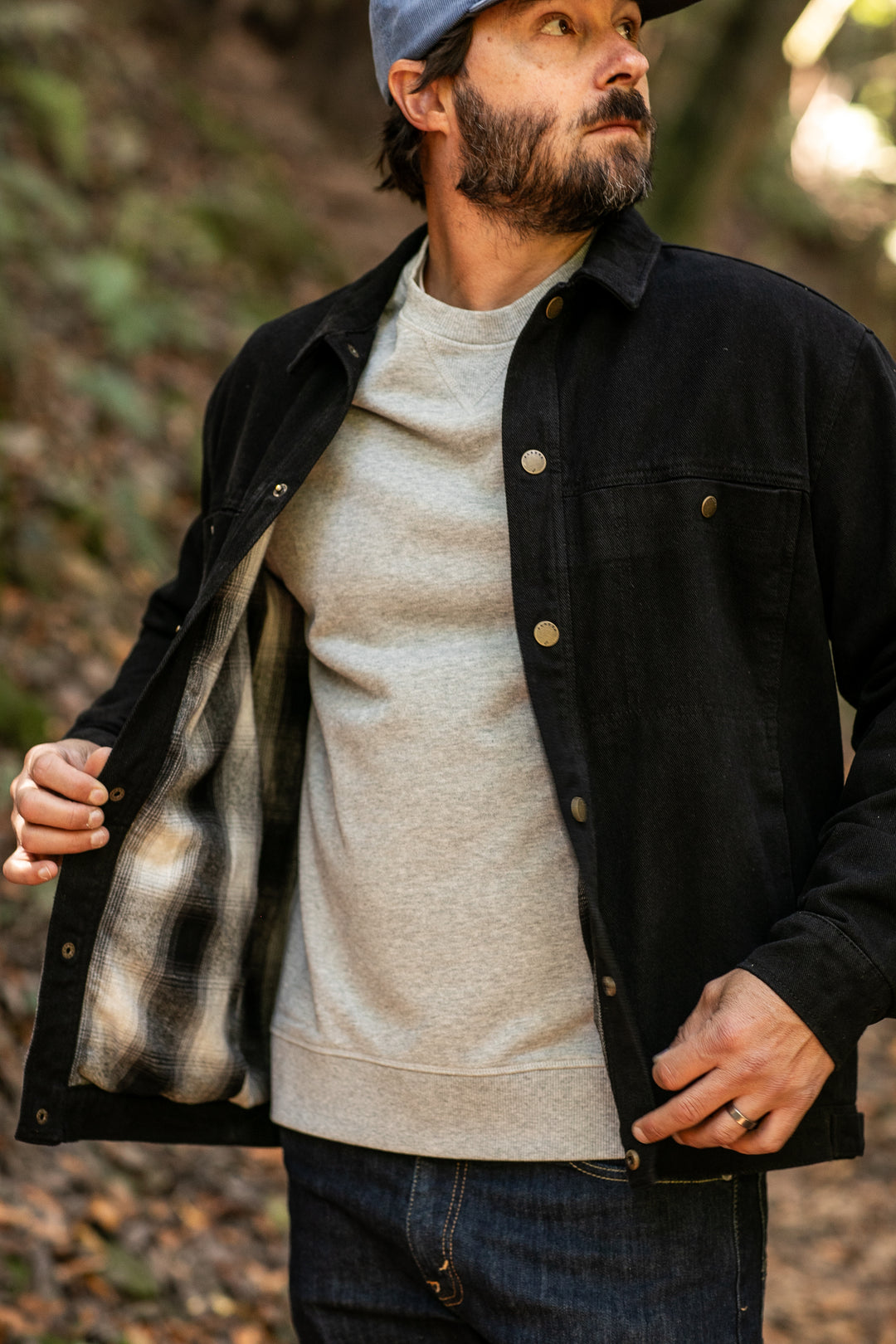 A bearded man in a gray cap and sweatshirt stands outdoors, showing the plaid flannel lining inside his open Men&