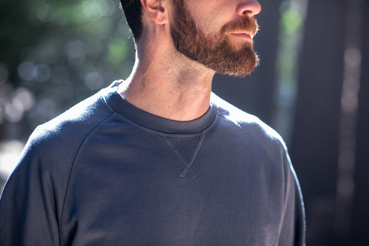 A bearded man wears the Pladra Men's Klamath Crew in Stone Blue while standing outdoors in soft sunlight.