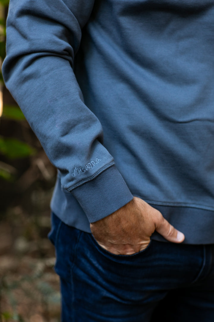 Close-up of a man's arm in Pladra Men's Klamath Crew - Stone Blue, logo on sleeve, hand in pocket.