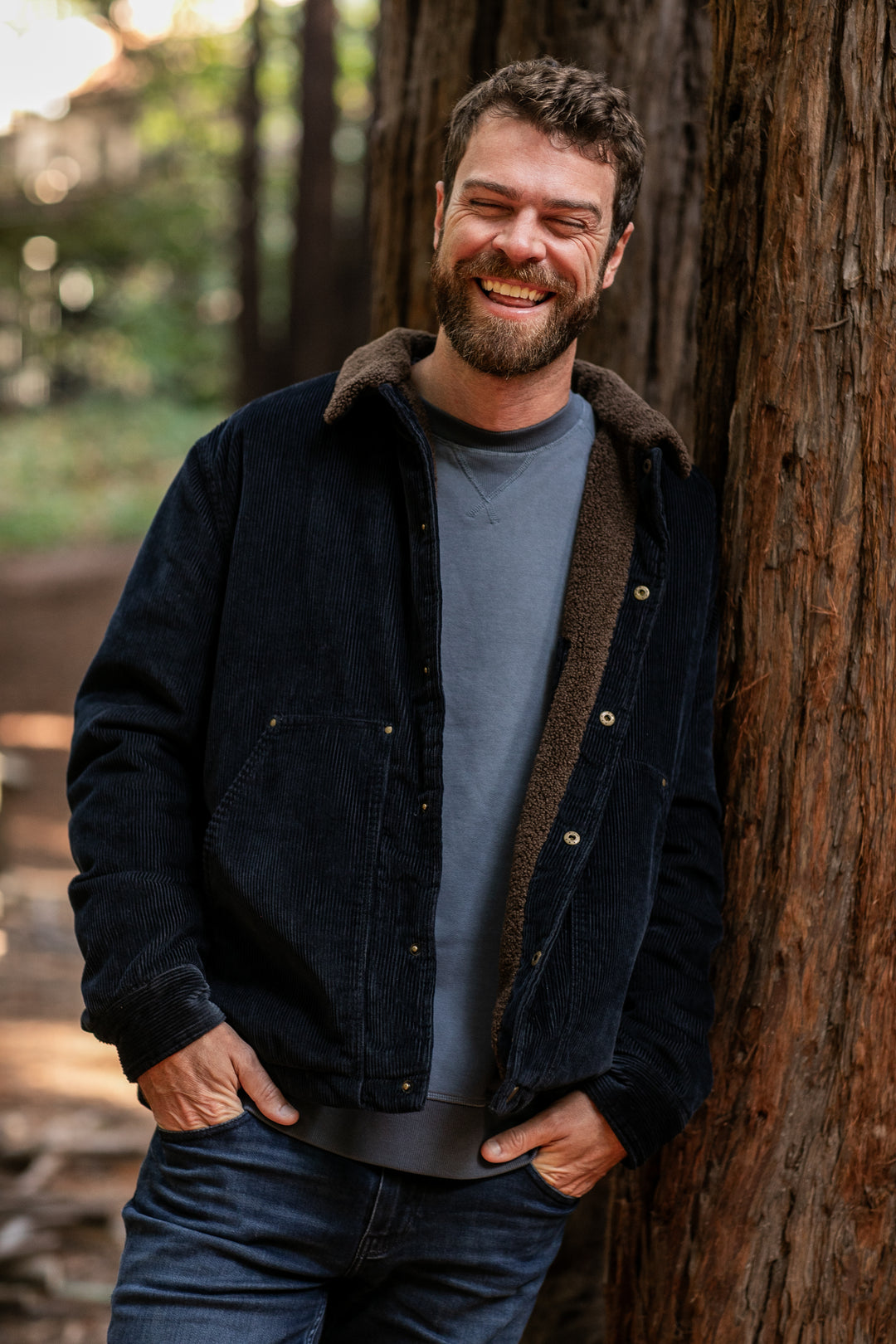 A bearded man, dressed in the Men's Ironwood Sherpa Jacket–Midnight Blue over a gray shirt and jeans, smiles warmly as he stands leaning against a tree outdoors with tall trees and soft natural light behind him.