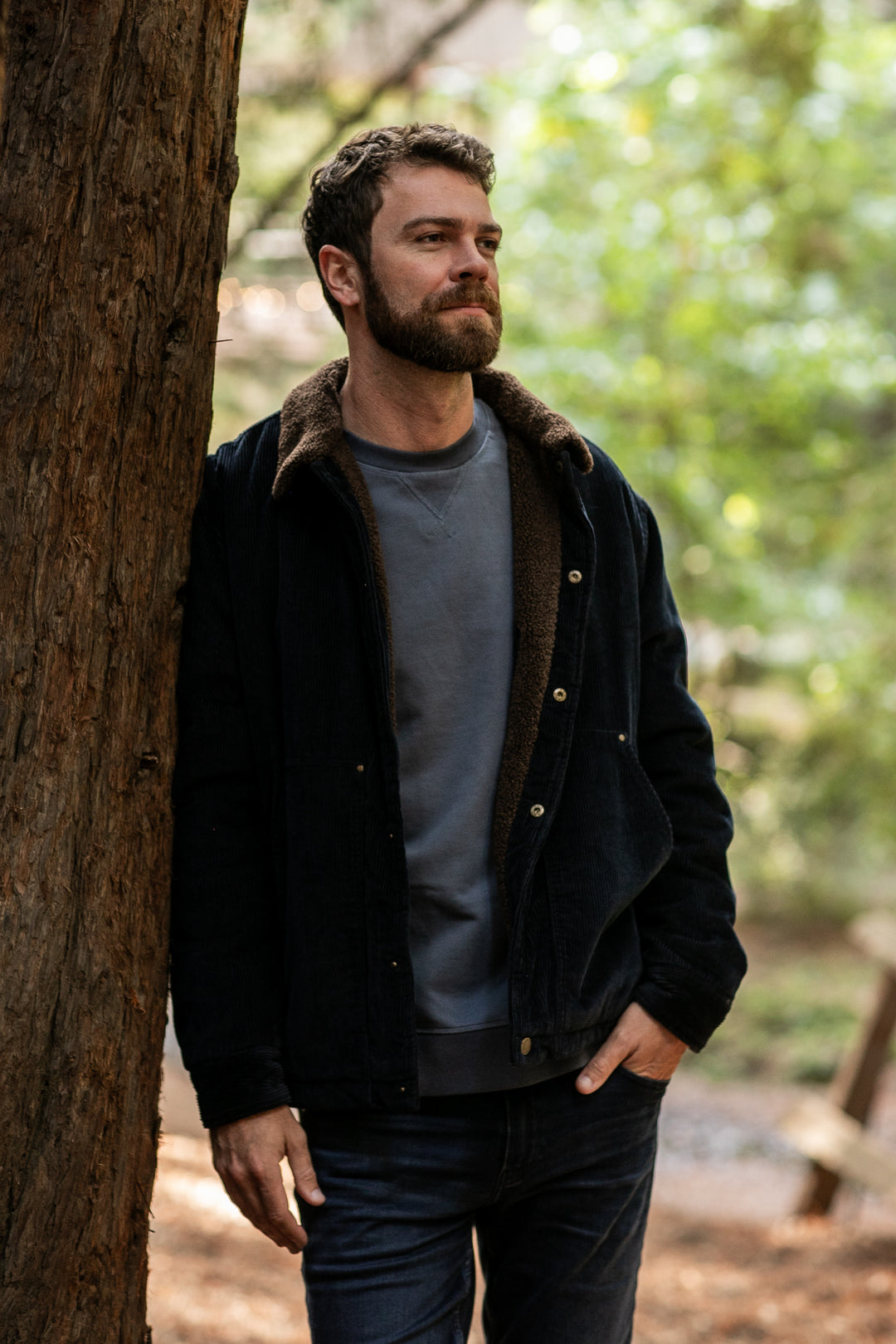 A bearded man stands outdoors, leaning against a tree and wearing the Men's Ironwood Sherpa Jacket in Midnight Blue over a gray sweater and jeans. Greenery and soft sunlight create a serene background.