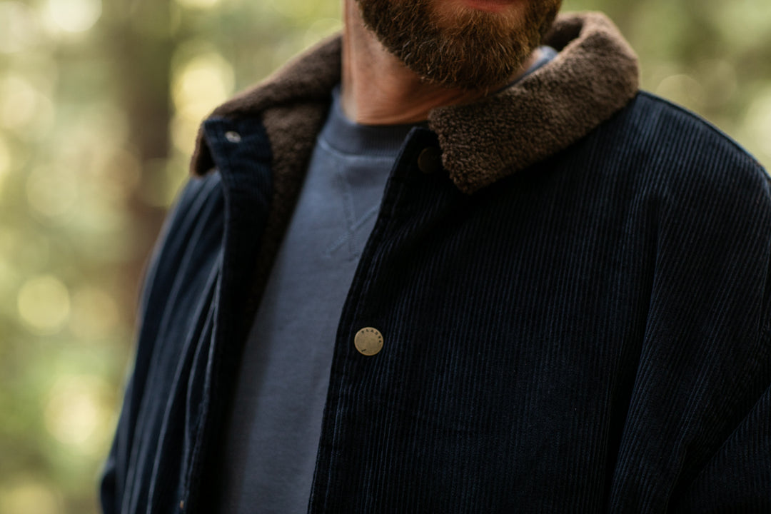 A close-up of a man wearing the Men's Ironwood Sherpa Jacket in Midnight Blue, featuring a dark cotton corduroy exterior, brown fleece collar, and blue shirt underneath, outdoors with blurred trees in the background.