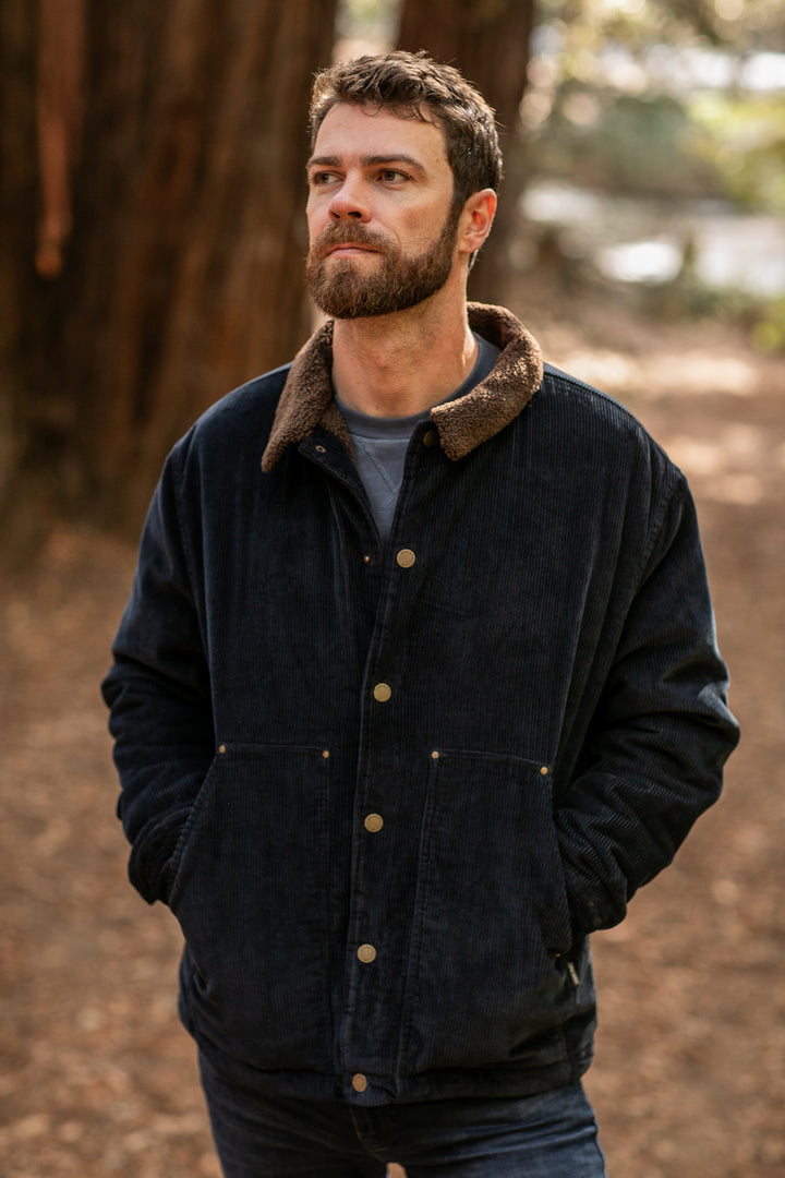 A bearded man stands outdoors in a wooded area, hands in pockets, wearing the Men's Ironwood Sherpa Jacket in Midnight Blue with a brown collar. Soft natural light highlights the cozy cotton corduroy fabric.