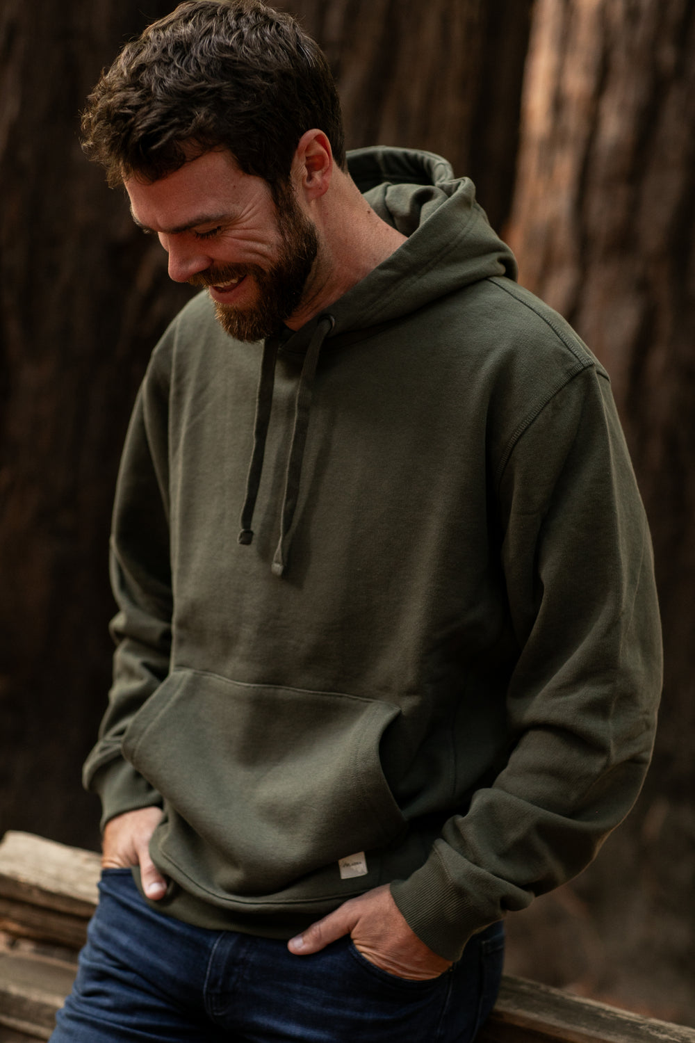 A bearded man, wearing the Men's Sierra Hoodie in Pine Green and blue jeans, stands outdoors with his hands in his pockets, smiling and looking down. Trees and wooden rails appear blurred in the background.