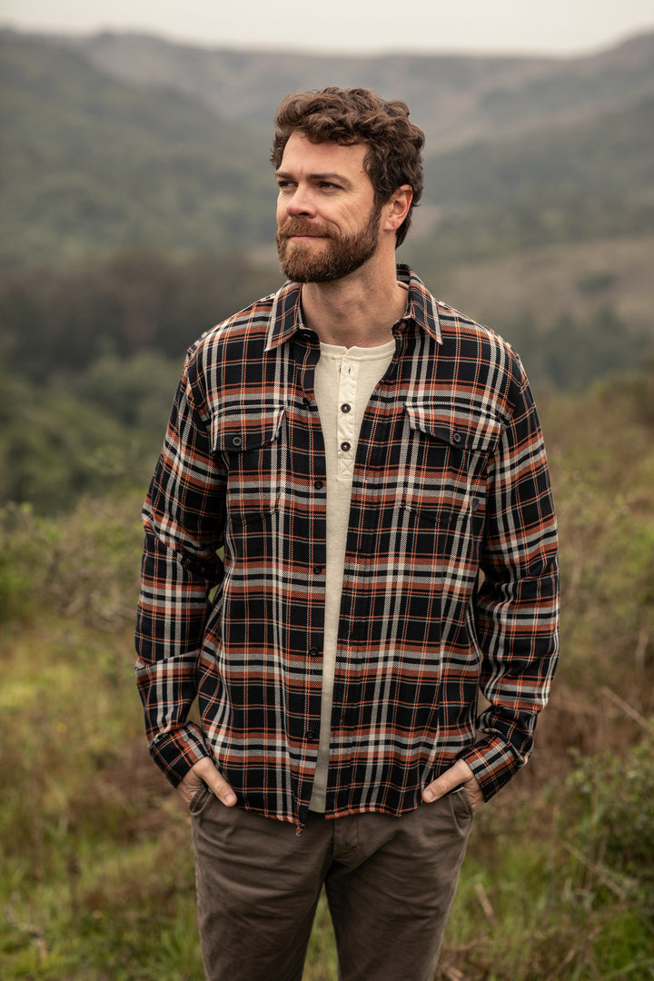 A bearded man in a Pladra Men’s Workhorse Elite Flannel Shirt stands outside before green hills, hands in pockets.