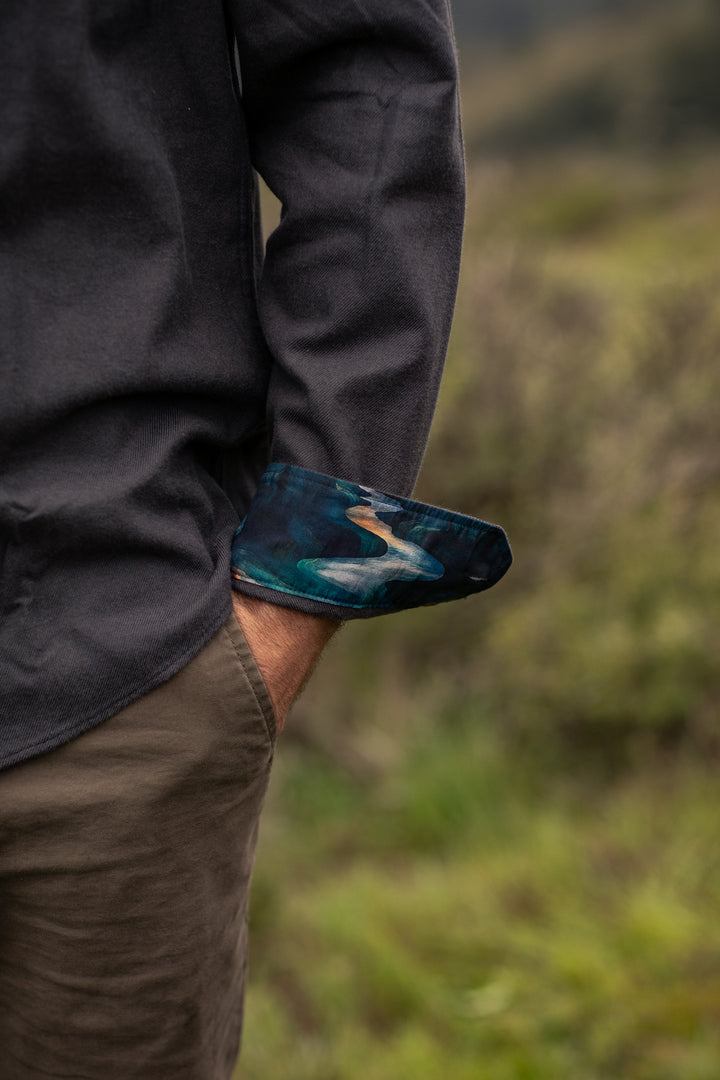 Person with one hand in pocket, wearing Pladra’s Men’s Cascade Flannel Shirt - Charcoal Grey and olive pants outdoors.