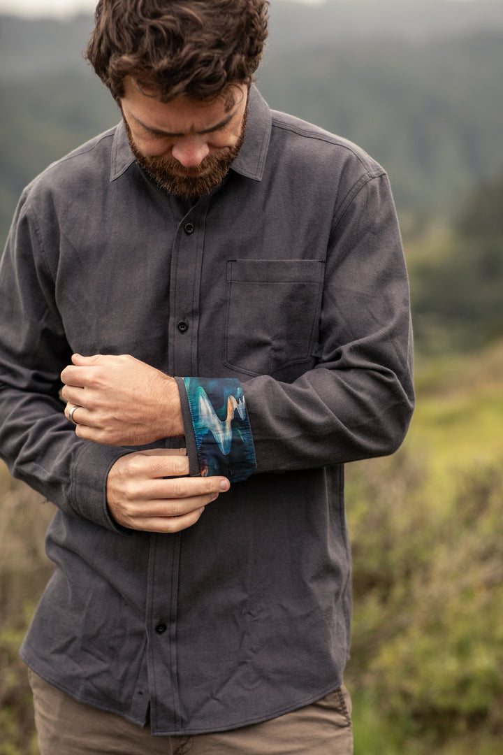 A man rolls up his sleeve outdoors, revealing the colorful cuff of a Pladra Men’s Cascade Flannel Shirt in Charcoal Grey.