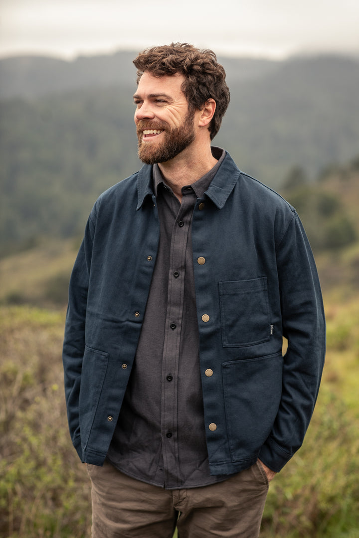 Smiling man outdoors in Pladra’s Men’s Cascade Flannel Shirt—Charcoal Grey, with blurred hills in the background.