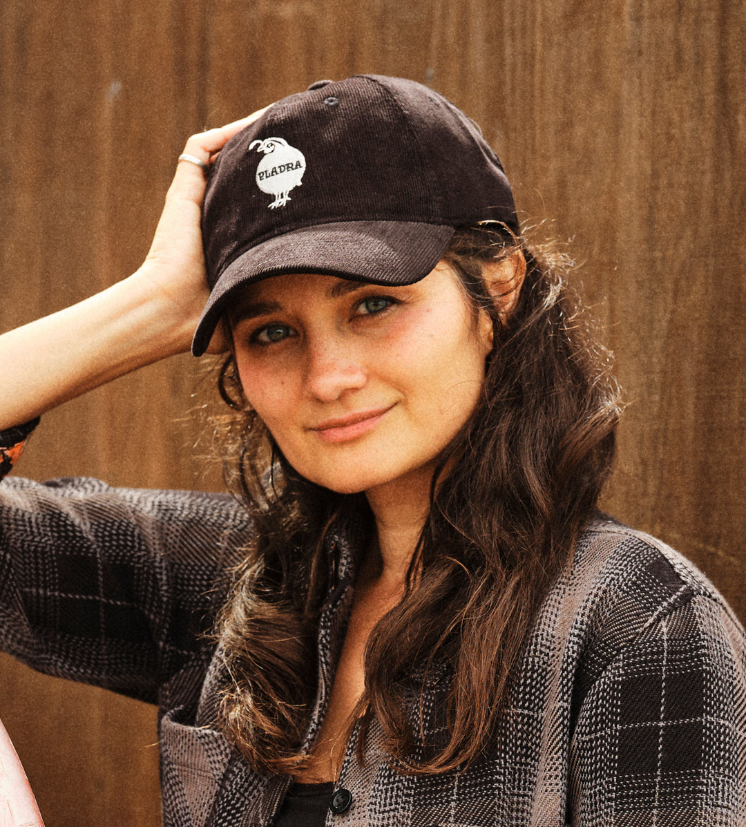 Woman in a plaid shirt wears the Pladra Quail Corduroy Dad Hat in black, smiling by a wooden background.