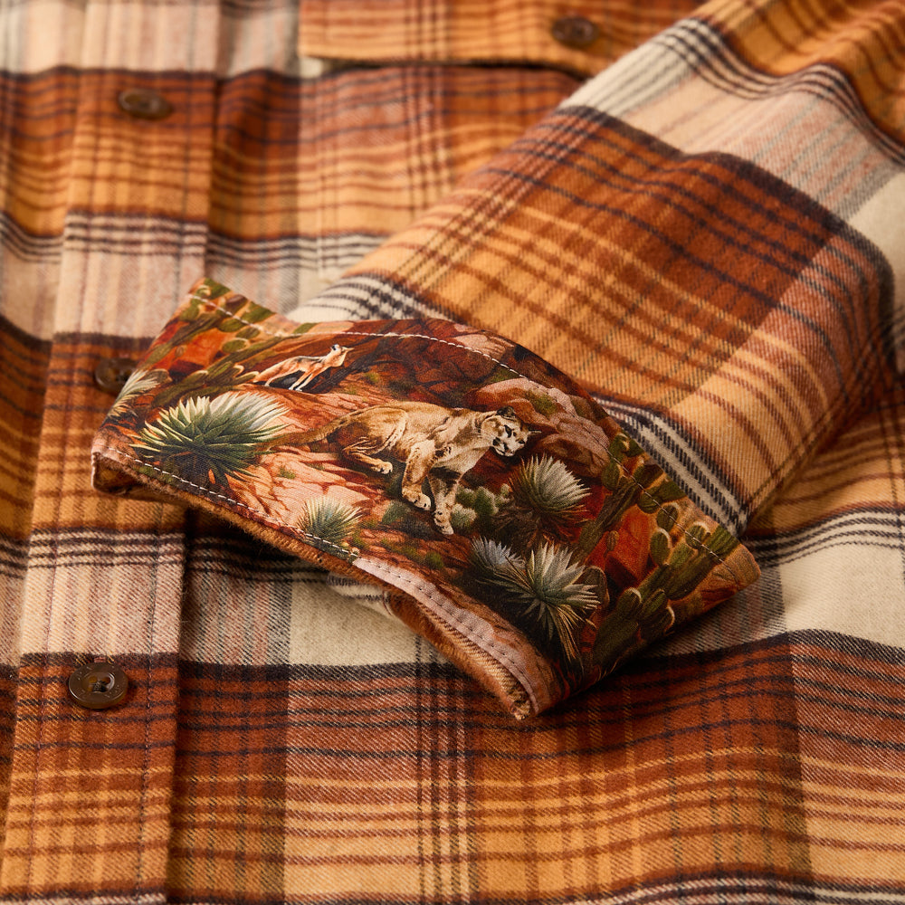 The Men's Workhorse Flannel Shirt in Sedona Orange showcases a brown, tan, and cream plaid in heavyweight cotton, with a cuff turned up to reveal a colorful desert scene lining featuring a mountain lion and cacti.