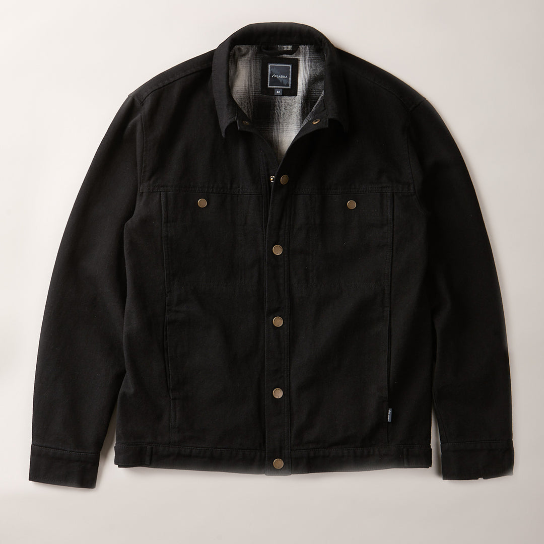 The Men's Sequoia Jacket- Shadow Black, made from black cotton twill with metal buttons, chest flap pockets, and side pockets, is laid flat on a light-colored surface.