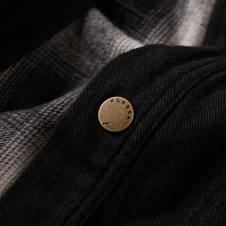A close-up of the metallic PLADRA button on the Men’s Sequoia Jacket- Shadow Black showcases the detailed texture of its black and grey plaid cotton twill fabric.