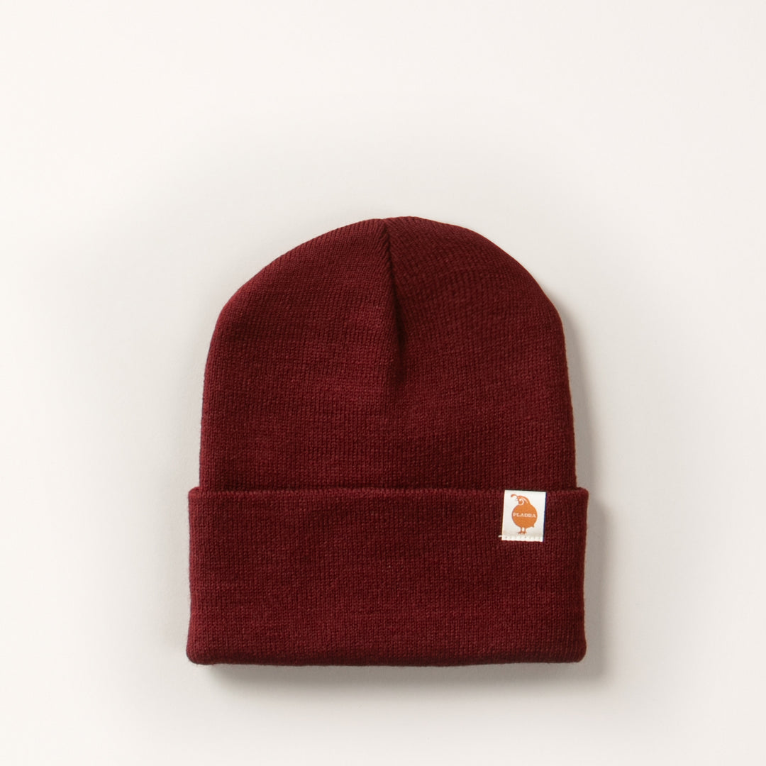The Stretch Cuff Beanie- Burgundy Red is a maroon acrylic beanie with a stretch cuff and a small white patch featuring a brown animal graphic on the front, shown against an off-white background.
