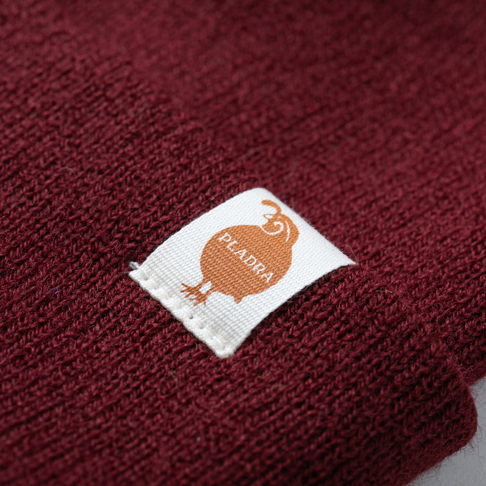 Close-up of the Stretch Cuff Beanie in Burgundy Red, featuring a white rectangular label with an orange bird and the word PEADRA.