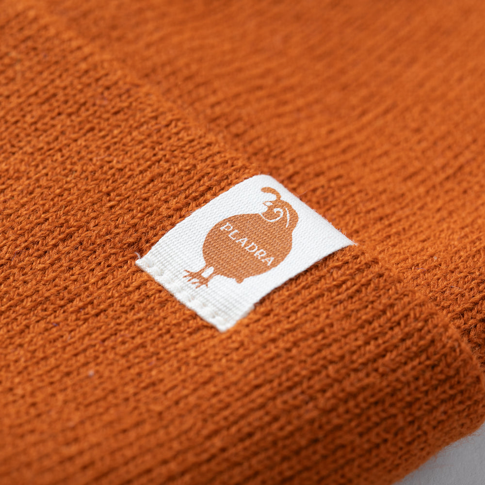 Close-up of Pladra's Stretch Cuff Beanie in burnt orange with white tag, brown bird logo, made in USA.
