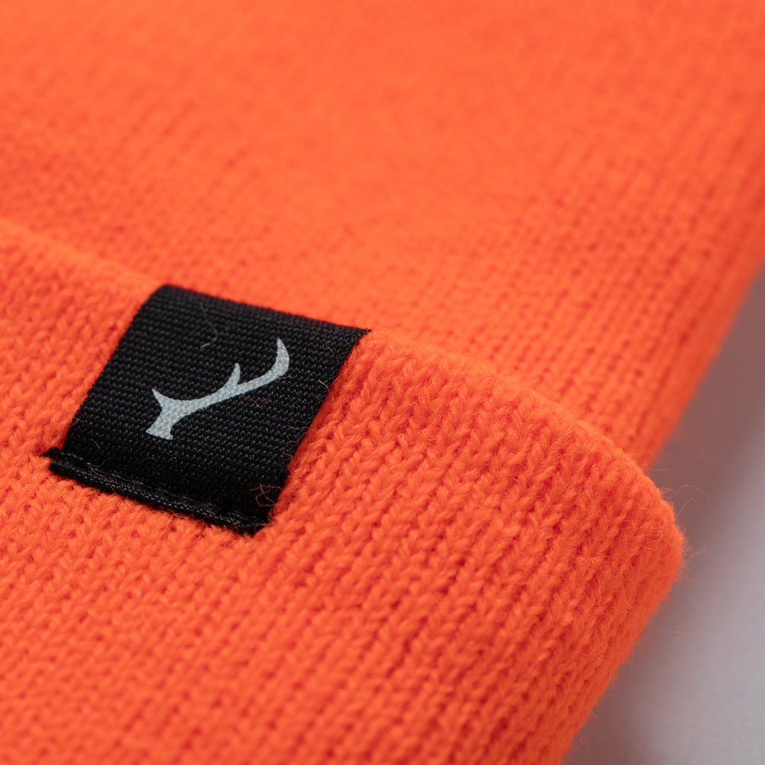 Close-up of the Stretch Cuff Beanie- Hunter Orange, showing its bright orange acrylic fabric and a black rectangular tag marked with a white antler-like symbol.