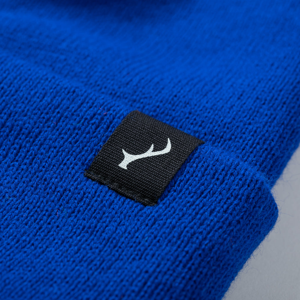 Close-up of Pladra Stretch Cuff Beanie- Royal Blue with a black label and white abstract symbol.