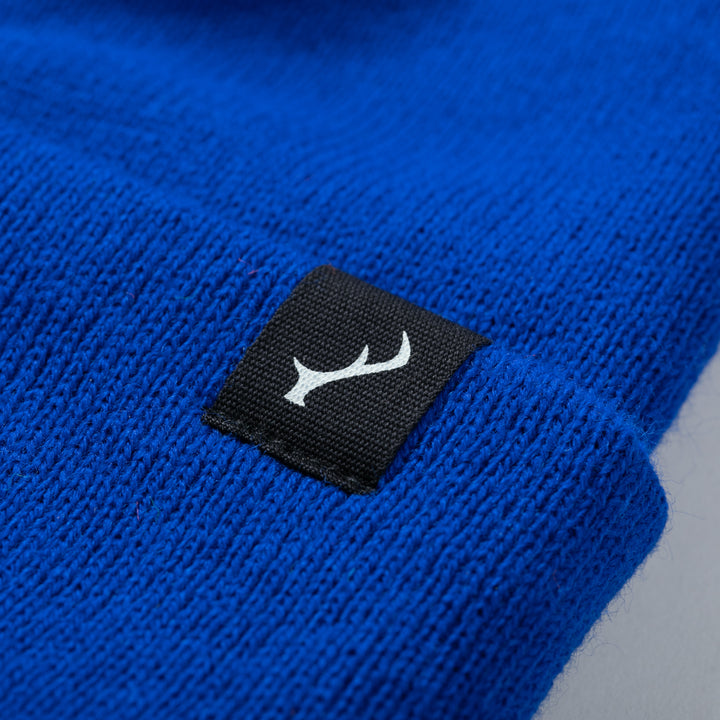 Close-up of Pladra Stretch Cuff Beanie- Royal Blue with a black label and white abstract symbol.