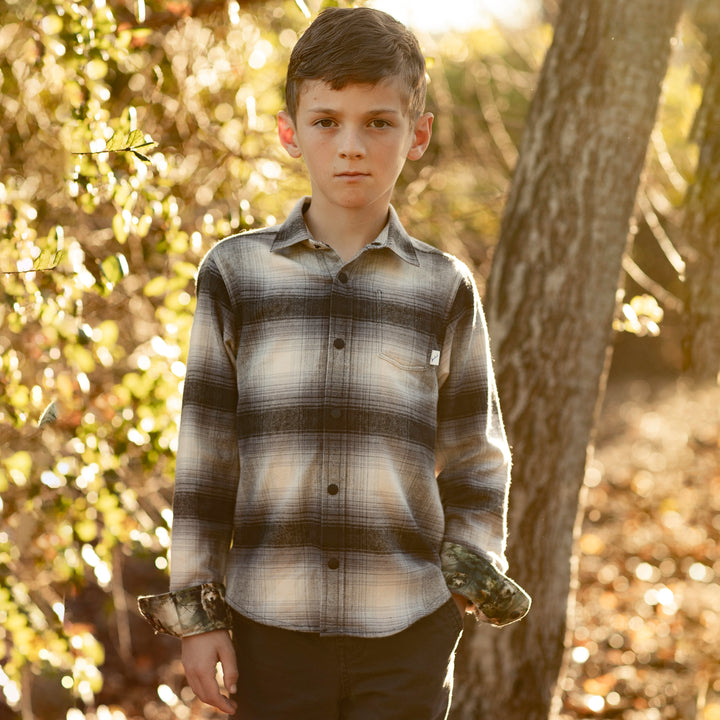 A boy wears the Pladra Toddler Cubs Flannel Shirt in Sterling Grey Heather outdoors among trees and sunlight.